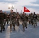 A large group of military personnel in camouflage uniforms are conducting a formation run or ruck march on a paved surface during early morning or dusk hours. A red guidon flag with a white crossed-keys or signal insignia is visible being carried mid-formation. The setting appears to be a military installation or base, with floodlights, fencing, and industrial structures visible in the background.