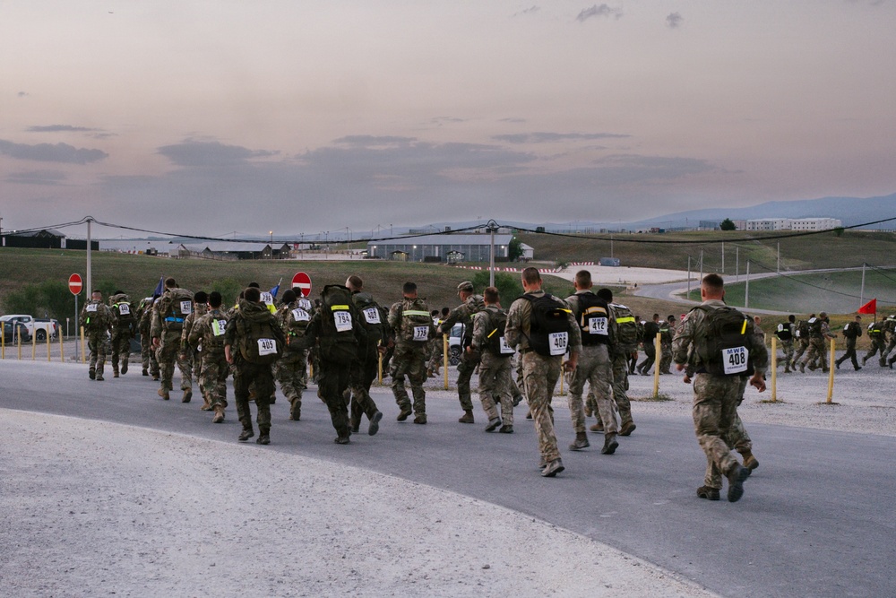A large group of military personnel in camouflage uniforms are conducting what appears to be a ruck march or military fitness event at dusk, moving along a paved road on what looks like a military base or installation. Participants are wearing numbered race bibs and high-visibility yellow reflective markers on their backs, along with loaded rucksacks. The setting appears to be a flat, open terrain with military infrastructure visible in the background.