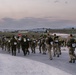 A large group of military personnel in camouflage uniforms are conducting what appears to be a ruck march or military fitness event at dusk, moving along a paved road on what looks like a military base or installation. Participants are wearing numbered race bibs and high-visibility yellow reflective markers on their backs, along with loaded rucksacks. The setting appears to be a flat, open terrain with military infrastructure visible in the background.