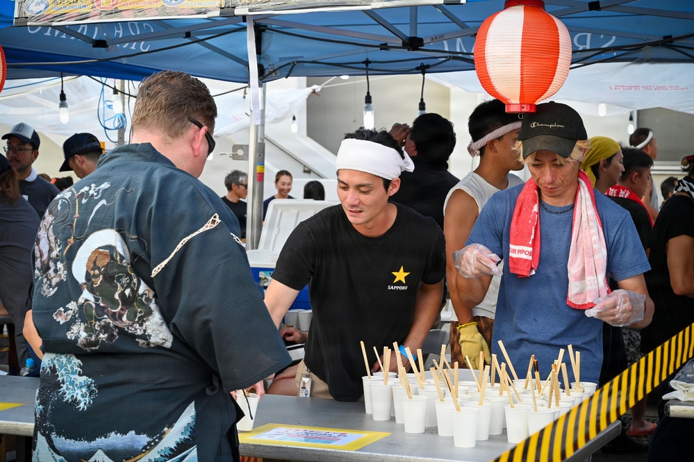 A Japanese food festival or outdoor market event where vendors are serving food from a booth. Two workers behind the counter are preparing and distributing small cups with chopsticks to customers. The setting features traditional Japanese decorative elements alongside modern branded apparel.