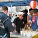 A Japanese food festival or outdoor market event where vendors are serving food from a booth. Two workers behind the counter are preparing and distributing small cups with chopsticks to customers. The setting features traditional Japanese decorative elements alongside modern branded apparel.