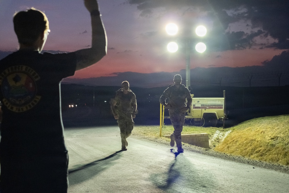 A nighttime outdoor scene at what appears to be a military installation, showing two individuals in camouflage uniforms running or walking along a paved road under bright portable light towers. A third person in a dark t-shirt is visible in the foreground with one arm raised, appearing to signal or direct the others. The scene is set against a twilight sky with a pink-red horizon.