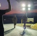 A nighttime outdoor scene at what appears to be a military installation, showing two individuals in camouflage uniforms running or walking along a paved road under bright portable light towers. A third person in a dark t-shirt is visible in the foreground with one arm raised, appearing to signal or direct the others. The scene is set against a twilight sky with a pink-red horizon.