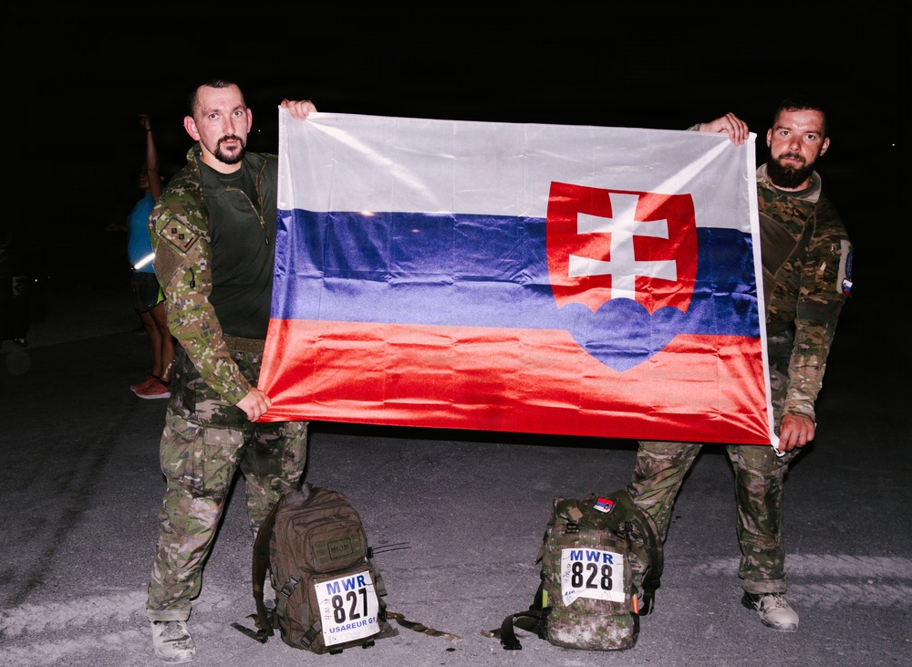 Two men in military camouflage uniforms are holding up a Slovak flag at night. They are standing behind backpacks labeled with MWR race numbers 827 and 828, indicating participation in a military ruck march or endurance event organized under USAREUR. The setting appears to be a paved outdoor area photographed under artificial lighting.