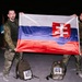 Two men in military camouflage uniforms are holding up a Slovak flag at night. They are standing behind backpacks labeled with MWR race numbers 827 and 828, indicating participation in a military ruck march or endurance event organized under USAREUR. The setting appears to be a paved outdoor area photographed under artificial lighting.
