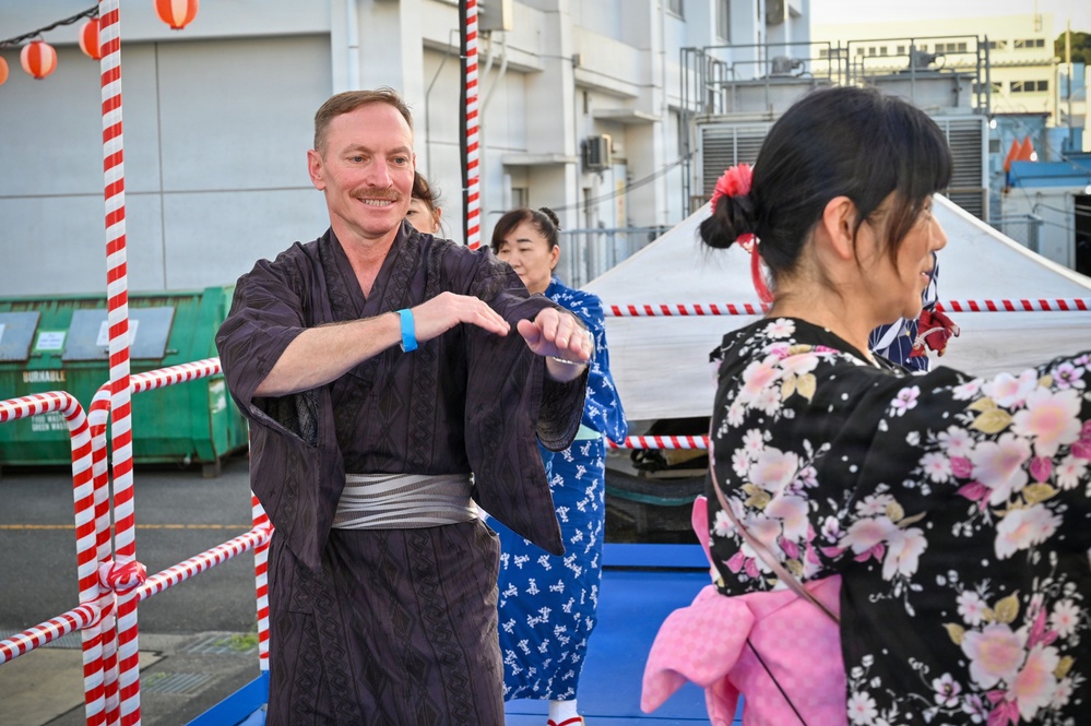 A group of people wearing traditional Japanese yukata (summer kimono) are participating in what appears to be a Bon Odori or similar Japanese festival dance. The setting is an outdoor area decorated with red and white rope barriers and paper lanterns, suggesting a matsuri (festival) event. A Western man in a dark yukata is smiling and mimicking dance movements, likely learning from the women around him.