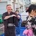 A group of people wearing traditional Japanese yukata (summer kimono) are participating in what appears to be a Bon Odori or similar Japanese festival dance. The setting is an outdoor area decorated with red and white rope barriers and paper lanterns, suggesting a matsuri (festival) event. A Western man in a dark yukata is smiling and mimicking dance movements, likely learning from the women around him.