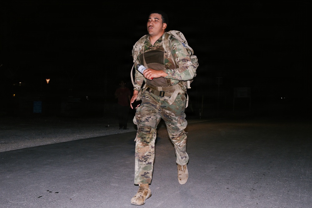 A soldier in full military gear is running or marching at night on a paved surface. The individual is wearing OCP (Operational Camouflage Pattern) uniform, a tactical vest/plate carrier, and is carrying a large rucksack. The scene appears to be a military training event or ruck march exercise conducted after dark.