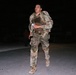 A soldier in full military gear is running or marching at night on a paved surface. The individual is wearing OCP (Operational Camouflage Pattern) uniform, a tactical vest/plate carrier, and is carrying a large rucksack. The scene appears to be a military training event or ruck march exercise conducted after dark.