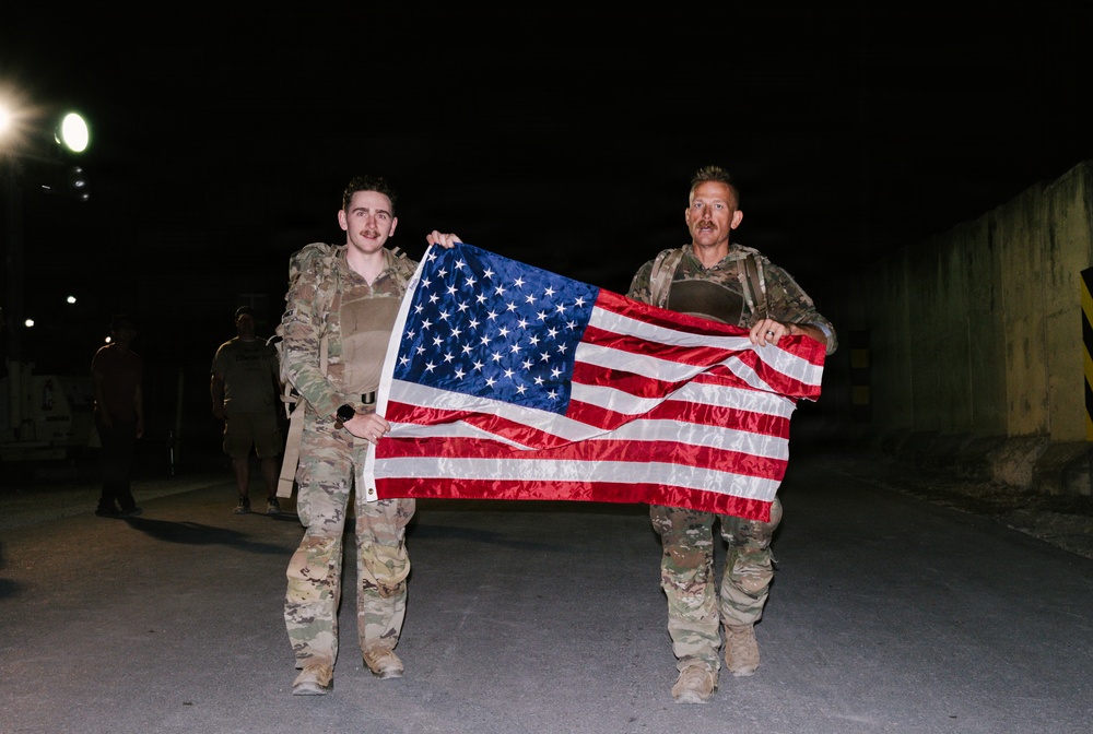 Two individuals in military camouflage uniforms are holding an American flag horizontally between them at night. They appear to be standing on a paved surface, possibly at a military installation or base, with additional personnel visible in the background. Both are wearing tactical gear including backpacks and body armor.
