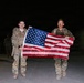 Two individuals in military camouflage uniforms are holding an American flag horizontally between them at night. They appear to be standing on a paved surface, possibly at a military installation or base, with additional personnel visible in the background. Both are wearing tactical gear including backpacks and body armor.