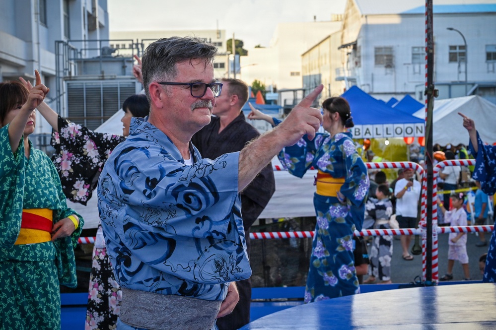 A group of people are participating in a traditional Japanese Bon Odori dance at an outdoor festival. The dancers are wearing yukata (summer kimono) and performing choreographed movements on a raised blue platform. The event appears to be a summer matsuri (festival) with vendor tents and spectators visible in the background.