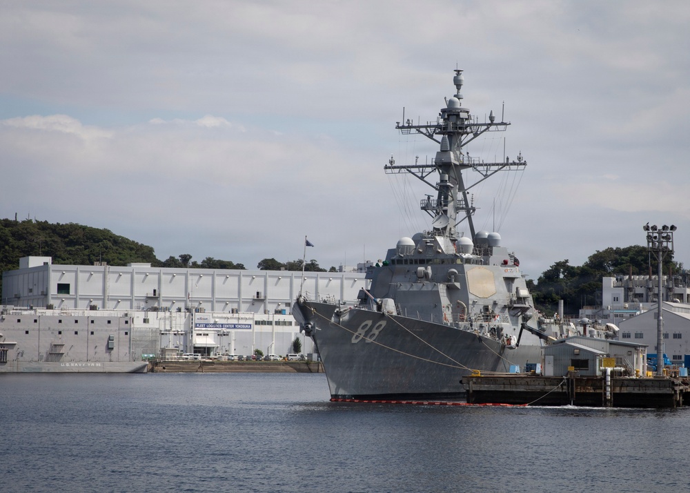 A U.S. Navy guided-missile destroyer bearing hull number 88 is moored at a naval installation identified as Fleet Logistics Center Yokosuka, Japan. The warship is docked alongside a pier with support facilities visible in the background. The scene depicts a routine port call or homeport berthing at a major U.S. naval base in the Pacific.