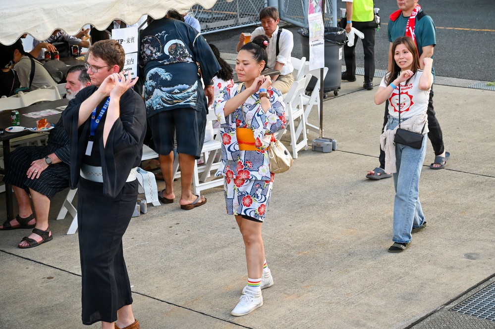 An outdoor festival or cultural event, likely in Japan, where attendees are participating in traditional Bon Odori-style dancing. Three people are prominently dancing in the foreground, including a woman wearing a yukata with floral patterns and an orange obi sash. The setting includes a tent with seating, Japanese signage, and a mix of Japanese and Western attendees.
