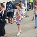 An outdoor festival or cultural event, likely in Japan, where attendees are participating in traditional Bon Odori-style dancing. Three people are prominently dancing in the foreground, including a woman wearing a yukata with floral patterns and an orange obi sash. The setting includes a tent with seating, Japanese signage, and a mix of Japanese and Western attendees.