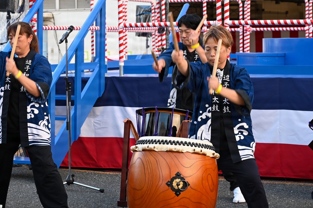Three performers dressed in matching dark blue happi coats with Japanese kanji text are playing taiko drums at what appears to be a Japanese festival or outdoor event. The central focus is a large barrel-shaped nagado-daiko drum with decorative rope lacing and a metal ornament. The background features red-and-white striped decorations and a blue stage structure draped with a red, white, and blue banner.