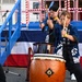 Three performers dressed in matching dark blue happi coats with Japanese kanji text are playing taiko drums at what appears to be a Japanese festival or outdoor event. The central focus is a large barrel-shaped nagado-daiko drum with decorative rope lacing and a metal ornament. The background features red-and-white striped decorations and a blue stage structure draped with a red, white, and blue banner.