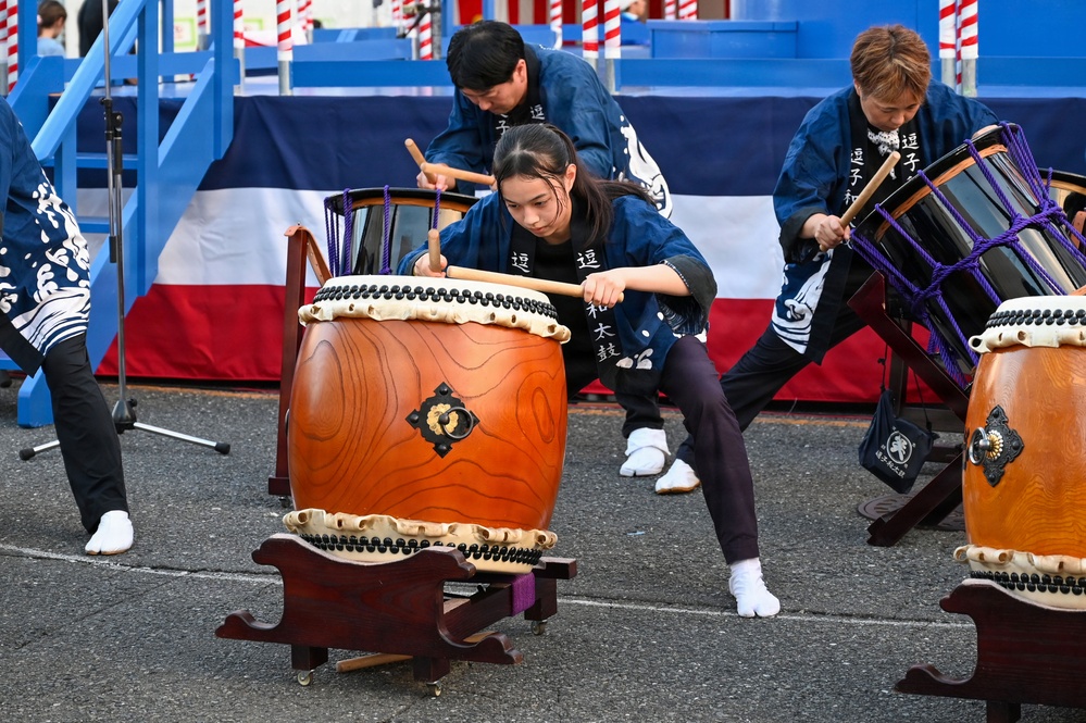 A group of performers are playing traditional Japanese taiko drums (和太鼓) outdoors at what appears to be a festival or public event. The performers are wearing matching blue happi coats with Japanese kanji characters, and are actively striking large barrel-shaped drums with wooden bachi sticks. The central focus is a young female drummer in a wide, low stance, demonstrating intense concentration and physical engagement.