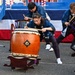 A group of performers are playing traditional Japanese taiko drums (和太鼓) outdoors at what appears to be a festival or public event. The performers are wearing matching blue happi coats with Japanese kanji characters, and are actively striking large barrel-shaped drums with wooden bachi sticks. The central focus is a young female drummer in a wide, low stance, demonstrating intense concentration and physical engagement.
