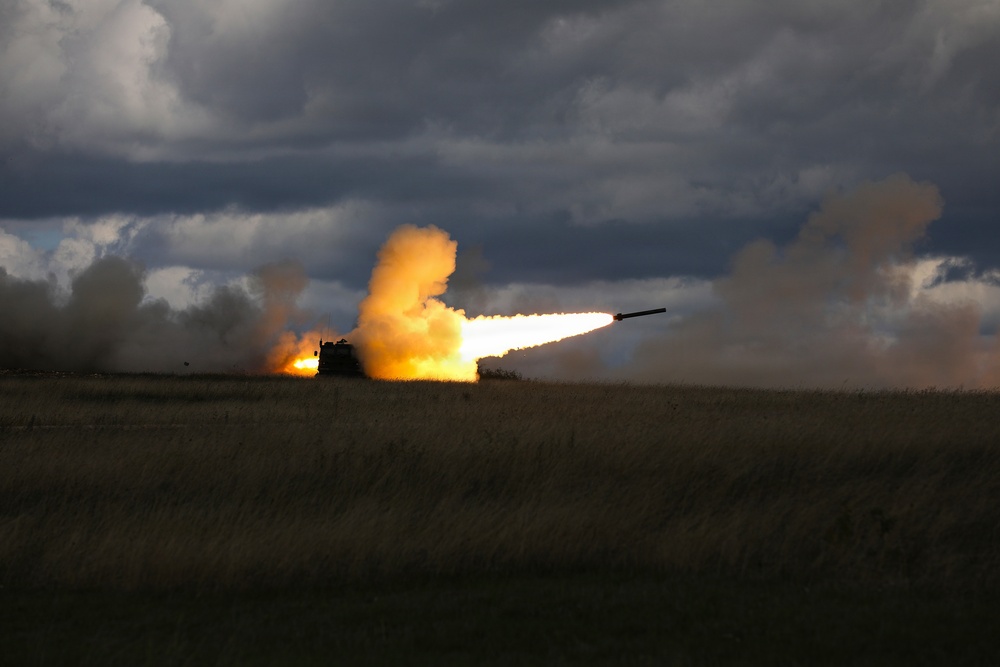 A military multiple rocket launch system (MLRS) is firing a rocket in an open field under a dramatic, overcast sky. The launch produces a large plume of orange and white smoke with a visible rocket in flight trailing flame. The vehicle is silhouetted against the intense light of the exhaust.