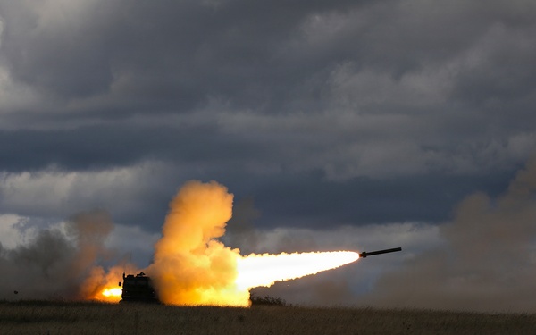 A military multiple rocket launch system (MLRS) is firing a rocket in an open field under a dramatic, overcast sky. The launch produces a large plume of orange and white smoke with a visible rocket in flight trailing flame. The vehicle is silhouetted against the intense light of the exhaust.