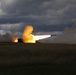 A military multiple rocket launch system (MLRS) is firing a rocket in an open field under a dramatic, overcast sky. The launch produces a large plume of orange and white smoke with a visible rocket in flight trailing flame. The vehicle is silhouetted against the intense light of the exhaust.