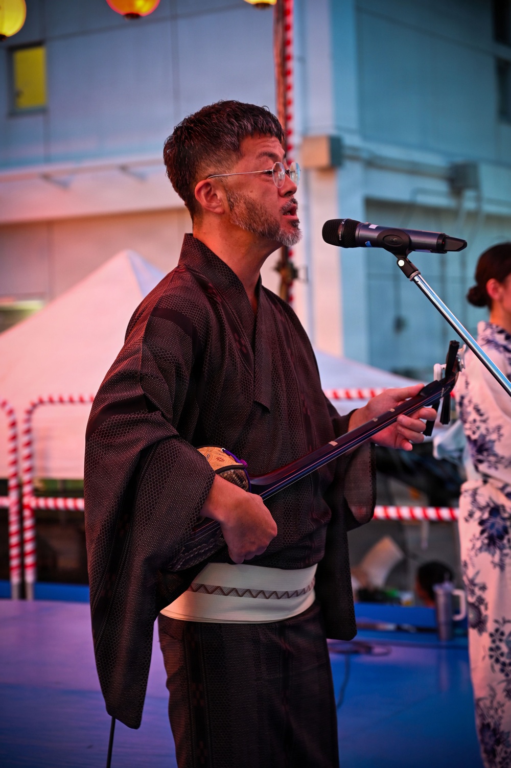A man wearing a dark traditional Japanese kimono with a white obi sash is performing at an outdoor event, singing into a microphone while holding what appears to be a shamisen (traditional Japanese three-stringed instrument). He wears round wire-framed glasses and has a short beard. The setting appears to be a Japanese festival or cultural event, evidenced by red and white decorations and paper lanterns visible in the background.