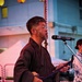 A man wearing a dark traditional Japanese kimono with a white obi sash is performing at an outdoor event, singing into a microphone while holding what appears to be a shamisen (traditional Japanese three-stringed instrument). He wears round wire-framed glasses and has a short beard. The setting appears to be a Japanese festival or cultural event, evidenced by red and white decorations and paper lanterns visible in the background.