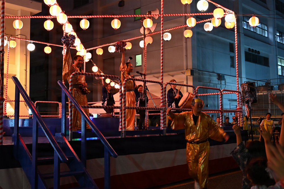 A nighttime Japanese festival (matsuri) scene featuring performers on an elevated stage decorated with traditional paper lanterns (chochin) and red-and-white striped rope barriers. Performers dressed in gold costumes are dancing and playing taiko drums on the stage, while a crowd gathers below. The festive atmosphere is enhanced by warm lantern lighting against a dark urban backdrop.