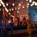 A nighttime Japanese festival (matsuri) scene featuring performers on an elevated stage decorated with traditional paper lanterns (chochin) and red-and-white striped rope barriers. Performers dressed in gold costumes are dancing and playing taiko drums on the stage, while a crowd gathers below. The festive atmosphere is enhanced by warm lantern lighting against a dark urban backdrop.