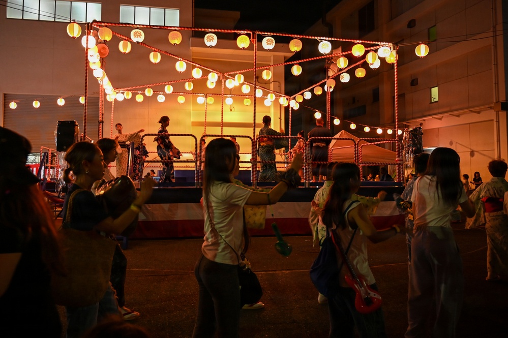 A traditional Japanese Bon Odori (Obon festival) dance event taking place at night in what appears to be a community or school courtyard. A central elevated yagura (wooden tower stage) is decorated with illuminated paper lanterns (chochin) and red-and-white rope trim, with performers dancing on top. Community members, some wearing yukata (summer kimono), circle the stage performing traditional folk dances.