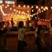 A traditional Japanese Bon Odori (Obon festival) dance event taking place at night in what appears to be a community or school courtyard. A central elevated yagura (wooden tower stage) is decorated with illuminated paper lanterns (chochin) and red-and-white rope trim, with performers dancing on top. Community members, some wearing yukata (summer kimono), circle the stage performing traditional folk dances.