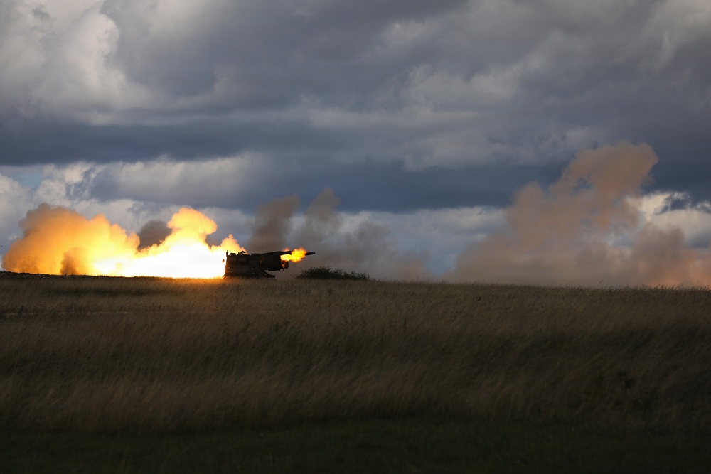 A large artillery piece or self-propelled howitzer is captured mid-fire on an open grassy plain, producing a massive muzzle blast with bright orange flames and billowing smoke clouds. The weapon appears to be firing horizontally across the landscape. The dramatic lighting and overcast sky with storm clouds create a striking visual contrast with the explosion.