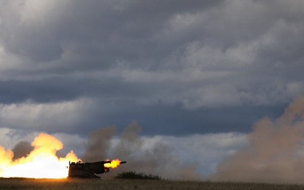 A large artillery piece or self-propelled howitzer is captured mid-fire on an open grassy plain, producing a massive muzzle blast with bright orange flames and billowing smoke clouds. The weapon appears to be firing horizontally across the landscape. The dramatic lighting and overcast sky with storm clouds create a striking visual contrast with the explosion.