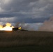 A large artillery piece or self-propelled howitzer is captured mid-fire on an open grassy plain, producing a massive muzzle blast with bright orange flames and billowing smoke clouds. The weapon appears to be firing horizontally across the landscape. The dramatic lighting and overcast sky with storm clouds create a striking visual contrast with the explosion.
