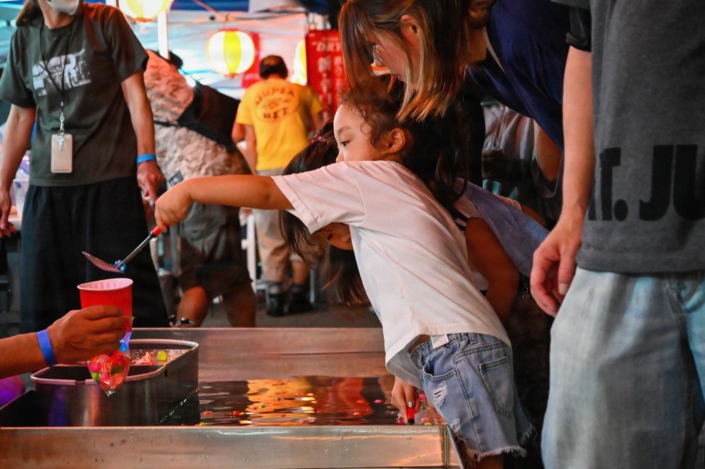 A young girl is participating in a traditional Japanese goldfish scooping game (kingyo-sukui) at what appears to be a summer festival (matsuri). She is leaning over a water tank using a small scoop (poi) to catch colorful items in the water, while an adult woman watches over her. The festive background includes paper lanterns and a crowd of festival-goers.