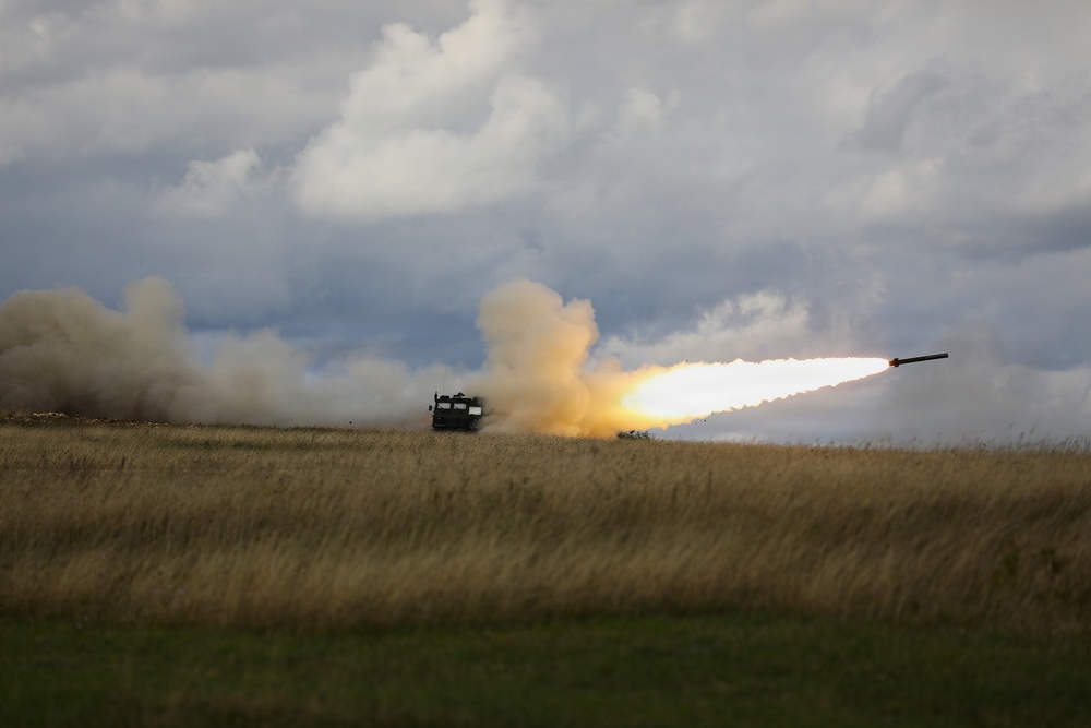 A military multiple rocket launch system (MLRS) is firing a rocket across an open grassy field under an overcast sky. A rocket is visibly in flight to the right with a bright flame trail, while large plumes of smoke billow from the launch vehicle. The setting appears to be a flat, rural or steppe-like terrain during daytime.