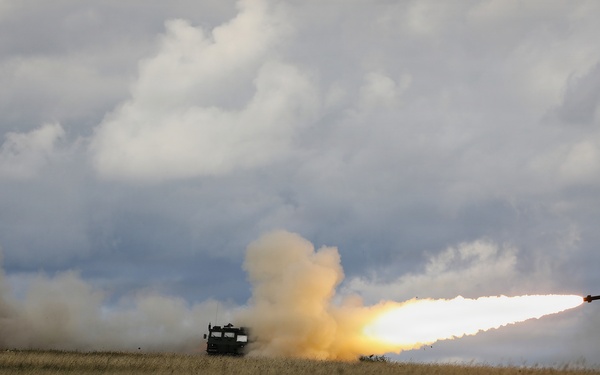 A military multiple rocket launch system (MLRS) is firing a rocket across an open grassy field under an overcast sky. A rocket is visibly in flight to the right with a bright flame trail, while large plumes of smoke billow from the launch vehicle. The setting appears to be a flat, rural or steppe-like terrain during daytime.