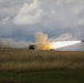A military multiple rocket launch system (MLRS) is firing a rocket across an open grassy field under an overcast sky. A rocket is visibly in flight to the right with a bright flame trail, while large plumes of smoke billow from the launch vehicle. The setting appears to be a flat, rural or steppe-like terrain during daytime.
