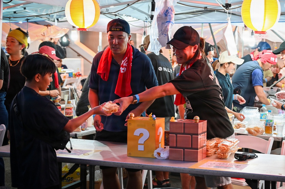 An outdoor festival or market event under a tent, where two adults wearing red scarves and baseball caps are handing food items to a child. The table features decorative boxes styled after Super Mario Bros. game elements, including a question mark block and a brick block. The setting appears to be an Asian cultural festival with paper lantern decorations and multiple vendors or activity stations in the background.