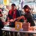 An outdoor festival or market event under a tent, where two adults wearing red scarves and baseball caps are handing food items to a child. The table features decorative boxes styled after Super Mario Bros. game elements, including a question mark block and a brick block. The setting appears to be an Asian cultural festival with paper lantern decorations and multiple vendors or activity stations in the background.