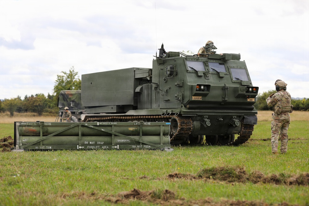 The image shows a tracked military rocket artillery system, identified as an M270 Multiple Launch Rocket System (MLRS), positioned in an open grassy field. Two soldiers in combat uniforms and helmets are visible near the vehicle, with one standing to the right observing and another working near the rear. A rocket pod reload container is positioned on the ground in the foreground with visible safety markings.