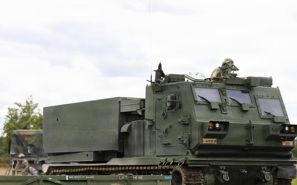 The image shows a tracked military rocket artillery system, identified as an M270 Multiple Launch Rocket System (MLRS), positioned in an open grassy field. Two soldiers in combat uniforms and helmets are visible near the vehicle, with one standing to the right observing and another working near the rear. A rocket pod reload container is positioned on the ground in the foreground with visible safety markings.