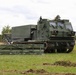The image shows a tracked military rocket artillery system, identified as an M270 Multiple Launch Rocket System (MLRS), positioned in an open grassy field. Two soldiers in combat uniforms and helmets are visible near the vehicle, with one standing to the right observing and another working near the rear. A rocket pod reload container is positioned on the ground in the foreground with visible safety markings.