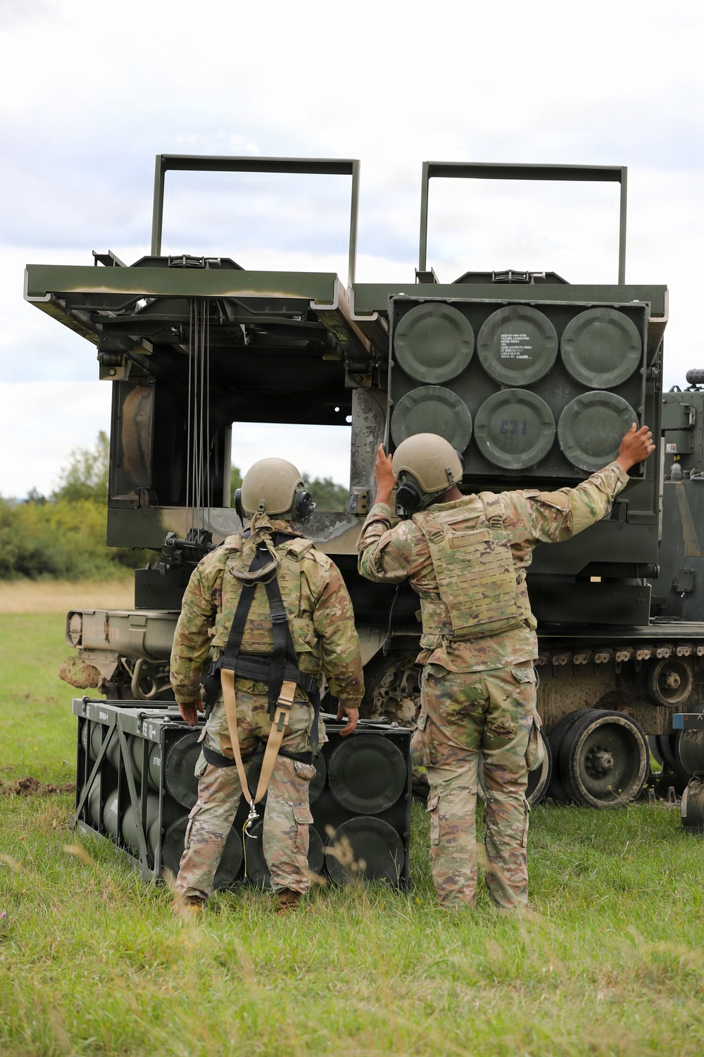 Two soldiers in U.S. Army MultiCam (OCP) uniforms are conducting a reloading operation on what appears to be an M270 Multiple Launch Rocket System (MLRS) or similar tracked rocket artillery platform. One soldier is handling a rocket pod/container while the other reaches toward the launcher assembly. The setting is an open grassy field with trees visible in the background under an overcast sky.