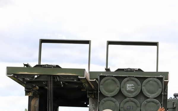 Two soldiers in U.S. Army MultiCam (OCP) uniforms are conducting a reloading operation on what appears to be an M270 Multiple Launch Rocket System (MLRS) or similar tracked rocket artillery platform. One soldier is handling a rocket pod/container while the other reaches toward the launcher assembly. The setting is an open grassy field with trees visible in the background under an overcast sky.