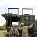 Two soldiers in U.S. Army MultiCam (OCP) uniforms are conducting a reloading operation on what appears to be an M270 Multiple Launch Rocket System (MLRS) or similar tracked rocket artillery platform. One soldier is handling a rocket pod/container while the other reaches toward the launcher assembly. The setting is an open grassy field with trees visible in the background under an overcast sky.