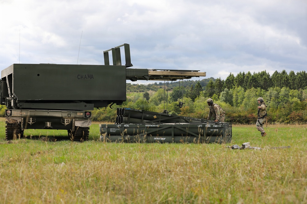 The image shows military personnel operating what appears to be an M270 Multiple Launch Rocket System (MLRS) or similar artillery rocket launcher in an open field. Two soldiers in combat uniforms are working with a rocket pod/container on the ground, likely during a reloading or maintenance operation. The setting appears to be a training exercise in a rural, wooded area under overcast skies.