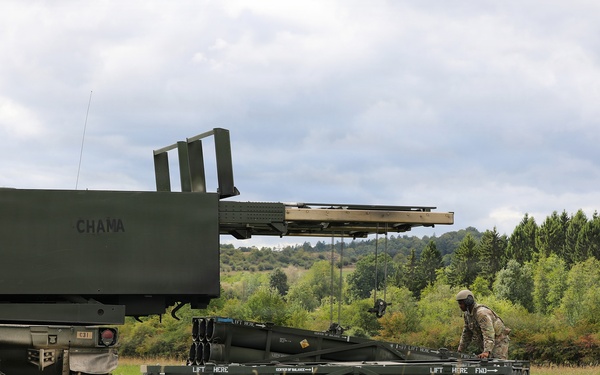 The image shows military personnel operating what appears to be an M270 Multiple Launch Rocket System (MLRS) or similar artillery rocket launcher in an open field. Two soldiers in combat uniforms are working with a rocket pod/container on the ground, likely during a reloading or maintenance operation. The setting appears to be a training exercise in a rural, wooded area under overcast skies.