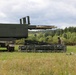 The image shows military personnel operating what appears to be an M270 Multiple Launch Rocket System (MLRS) or similar artillery rocket launcher in an open field. Two soldiers in combat uniforms are working with a rocket pod/container on the ground, likely during a reloading or maintenance operation. The setting appears to be a training exercise in a rural, wooded area under overcast skies.